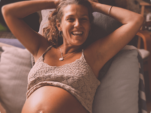 Pregnant woman smiling, with her arms behind her head and top raise to reveal her belly.