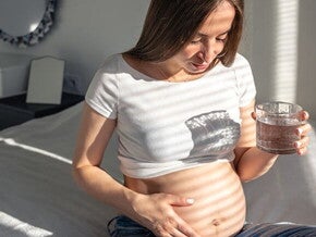 Gestante sentada na cama com uma mão sobre a barriga segurando um copo d’água