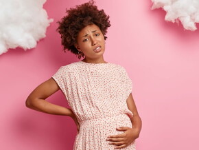 Mulher grávida em frente a uma parede rosa passando a mão na barriga e com expressão de desconforto. Mulher grávida em frente a uma parede rosa passando a mão na barriga e com expressão de desconforto.