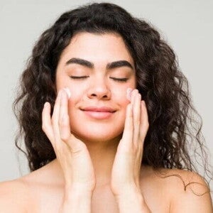 Mulher espalhando creme hidratante no rosto, de olhos fechados