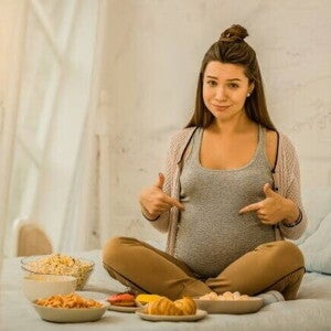 grávida aponta para a barriga sentada em uma cama, rodeada de pratos variados. grávida aponta para a barriga sentada em uma cama, rodeada de pratos variados.