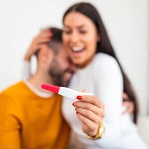 Mulher feliz segurando um teste de gravidez na mão e sendo abraçada por um homem, sugerindo comemoração pelo resultado positivo Mulher feliz segurando um teste de gravidez na mão e sendo abraçada por um homem, sugerindo comemoração pelo resultado positivo