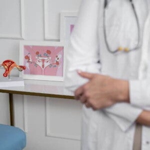 Médica com jaleco de branco ao lado de modelos anatômicos de órgãos reprodutivos femininos.