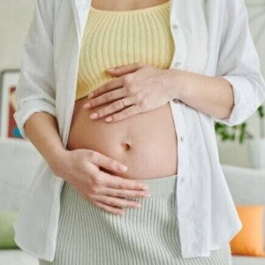 Uma gestante em pé em uma sala, com as mãos sobre a barriga.