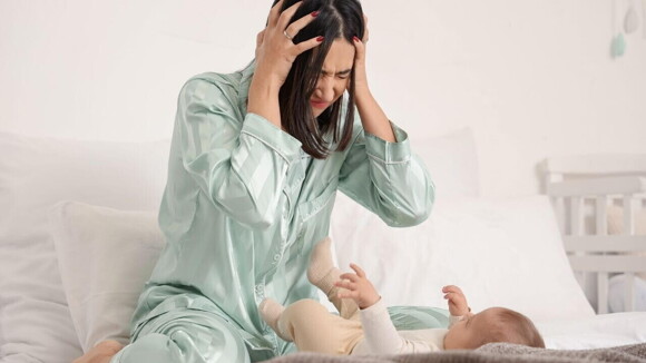 Uma mãe com as mãos na cabeça e aspecto de desespero, sentada na cama, em frente de um bebê deitado. Uma mãe com as mãos na cabeça e aspecto de desespero, sentada na cama, em frente de um bebê deitado.