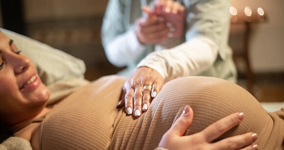 Mulher grávida deitada em uma cama recebendo o toque suave de uma doula em sua barriga Mulher grávida deitada em uma cama recebendo o toque suave de uma doula em sua barriga