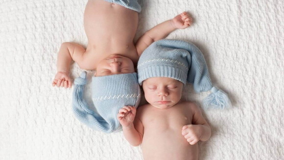 Dois bebês deitados lado a lado em uma cama, vestindo toucas iguais.