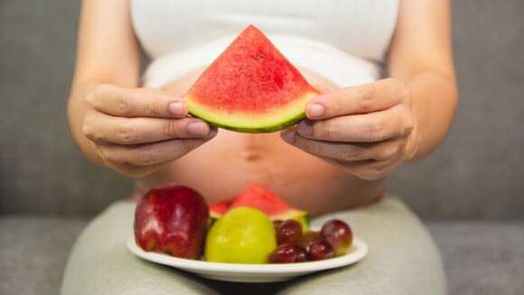 Mulher grávida com um prato de frutas no colo segurando uma fatia de melancia Mulher grávida com um prato de frutas no colo segurando uma fatia de melancia