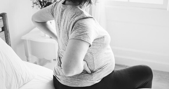 Grávida sentada na cama com as mãos apoiadas nas costas Grávida sentada na cama com as mãos apoiadas nas costas