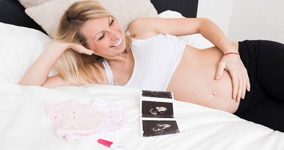 Mulher deitada em uma cama e sorrindo, com a mão na barriga, ao lado de um exame de ultrassom, um teste de gravidez e uma roupinha de bebê. Mulher deitada em uma cama e sorrindo, com a mão na barriga, ao lado de um exame de ultrassom, um teste de gravidez e uma roupinha de bebê.