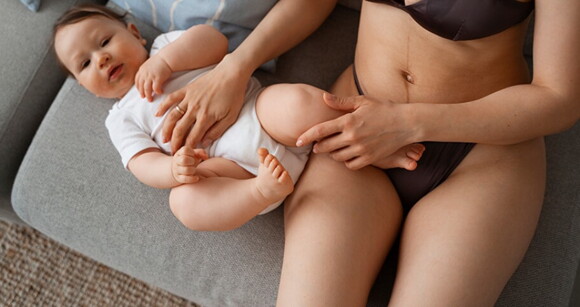 Mãe sentada no sofá ao lado do seu bebê usufruindo do conforto da calcinha pós-parto Mãe sentada no sofá ao lado do seu bebê usufruindo do conforto da calcinha pós-parto