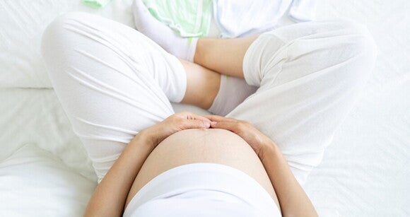 mulher com roupa branca sentada na cama enquanto segura a barriga de grávida mulher com roupa branca sentada na cama enquanto segura a barriga de grávida