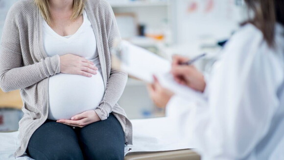 Uma gestante sentada com as mãos na barriga, sendo avaliada por uma profissional da saúde. Uma gestante sentada com as mãos na barriga, sendo avaliada por uma profissional da saúde.